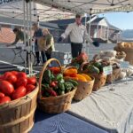 A volunteer stands with his arms open behind a table filled with brightly colored fall veggies.